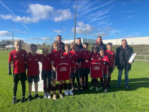 Fußballmannschaft in roten Trikots steht auf einem Rasenplatz, lächelt in die Kamera.