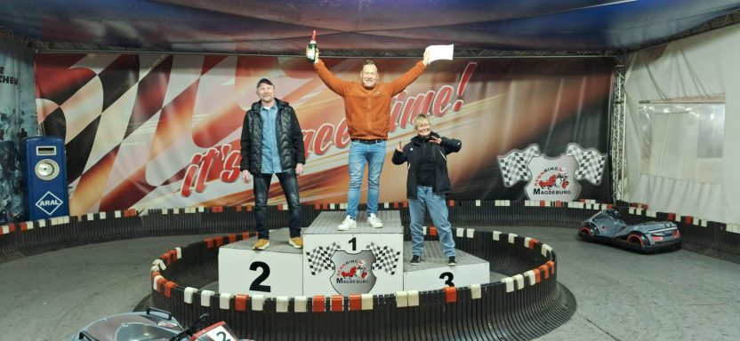 Sieger auf dem Podium, zwei Personen daneben, Rennstrecke im Hintergrund.